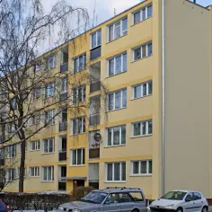 Żółty blok mieszkalny z oknami i drzewem przed nim.